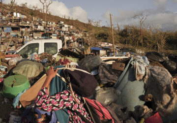 A woman carrying her belongings walks past debris after Cyclone Chido in the Kaweni slum Thursday, Dec. 19, 2024, on the outskirts of Mamoudzou, in the French Indian Ocean island of Mayotte. (AP Photo/Adrienne Surprenant)