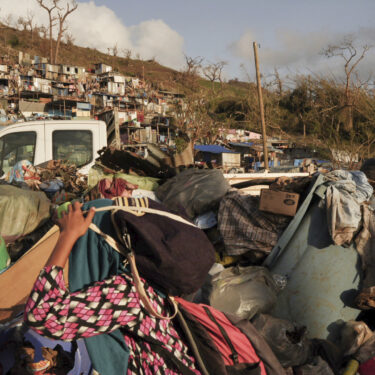 A woman carrying her belongings walks past debris after Cyclone Chido in the Kaweni slum Thursday, Dec. 19, 2024, on the outskirts of Mamoudzou, in the French Indian Ocean island of Mayotte. (AP Photo/Adrienne Surprenant)